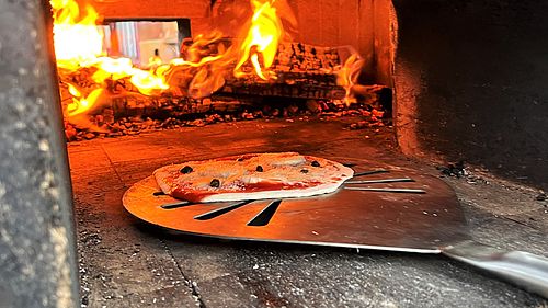 Im Backwagen des Vereins „Bündnis Feuer & Flamme“ brennt ein Feuer, im Vordergrung wird eine Pizza auf einem Pizzaschieber hineingeschoben.