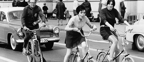 This black-and-white photo shows a busy city street. In the foreground on the right, two women are riding folding bikes. Riding behind them is a man on a regular men’s bicycle. An automobile can be seen next to the three cyclists along with another one behind them. In the background, there is a streetcar and several passengers. 
