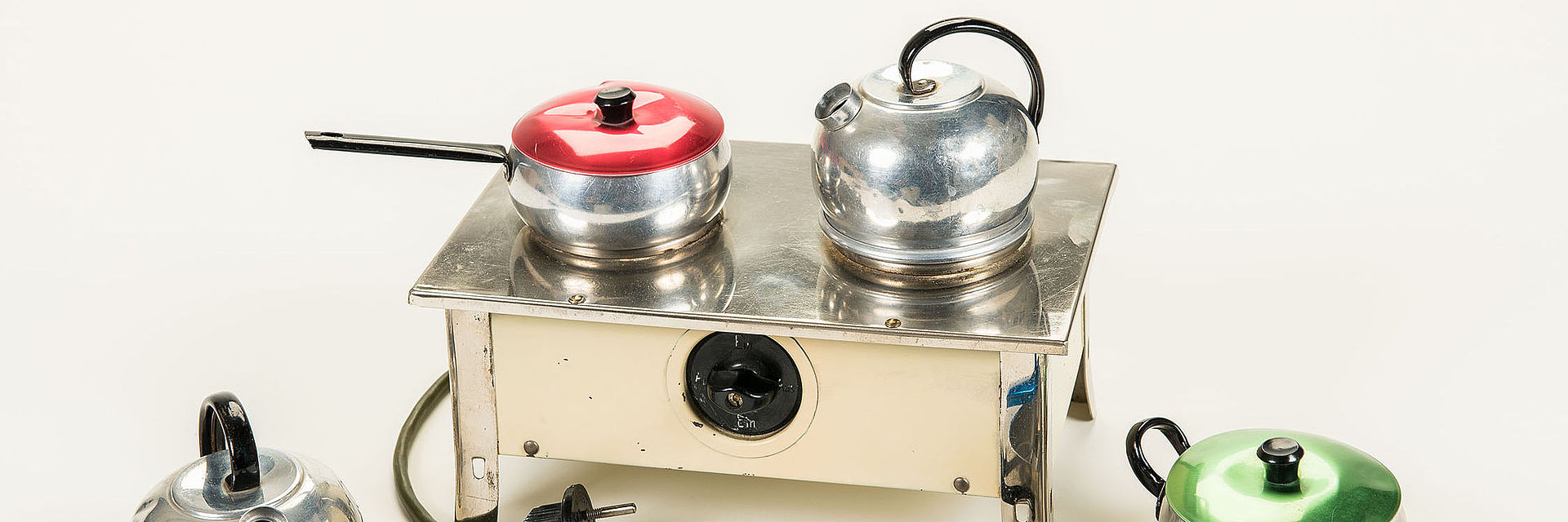 Dieser hell-emaillierte Miniaturherd wird elektrisch betrieben. Ausgestattet mit einem Netzkabel und Stecker, wird der Herd an der Vorderseite über einen Drehschalter ein- und ausgeschaltet. Auf zwei Kochplatten wurde mit dem dazugehörigen metallenen Kochgeschirr - zwei Wasserkessel, ein Stielkochtopf mit rotem Deckel und zwei Kochtöpfe mit einem blauen und einem grünen Deckel - gekocht. 