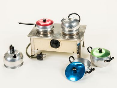 This brightly enameled miniature stove is operated electrically. The stove is equipped with a power cord and plug and has a rotary switch at the front for turning it on and off. Cooking was carried out on two hotplates with the accompanying metal cookware: two kettles, a saucepan with a red lid and two cooking pots with a blue and a green lid, respectively.