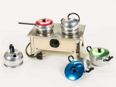 Dieser hell-emaillierte Miniaturherd wird elektrisch betrieben. Ausgestattet mit einem Netzkabel und Stecker, wird der Herd an der Vorderseite über einen Drehschalter ein- und ausgeschaltet. Auf zwei Kochplatten wurde mit dem dazugehörigen metallenen Kochgeschirr - zwei Wasserkessel, ein Stielkochtopf mit rotem Deckel und zwei Kochtöpfe mit einem blauen und einem grünen Deckel - gekocht. 