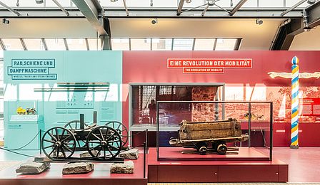 The picture on the right shows a cultural-historical exhibition unit of the Rail Transport exhibition entitled “A revolution in mobility” on a wine-red surface. On the left, an exhibition unit entitled “Wheel, rail and steam engine” is shown in front of a turquoise-colored surface explaining technical principles. In the foreground, two objects in display cases.
