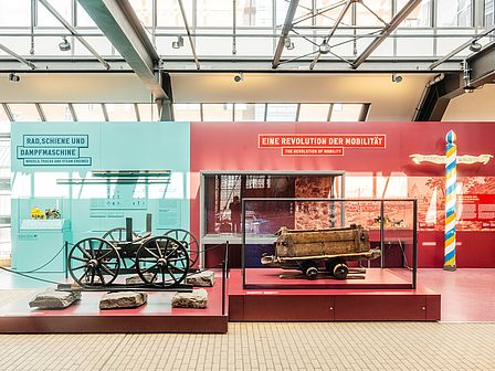 Rechts im Bild ist eine kulturgeschichtlich orientierte Ausstellungseinheit der Ausstellung Schienenverkehr mit der Überschrift „Eine Revolution der Mobilität“ auf einer weinroten Fläche zu sehen. Links wird eine Ausstellungseinheit mit der Überschrift „Rad, Schiene und Dampfmaschine“ vor einer türkisfarbenen Fläche gezeigt, die technische Prinzipien erklärt. Im Vordergrund zwei Objekte in Vitrinen.