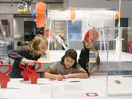 Drei Kinder stehen an einem Ausstellungstisch und tragen Daten in ein Heft ein.