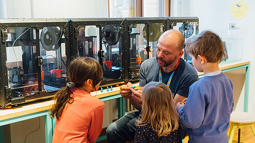 Ein Mann und drei Kinder sitzen vor einer Reihe von 3D-Druckern. Der Mann erklärt etwas, die Kinder haben Spaß und hören zu.