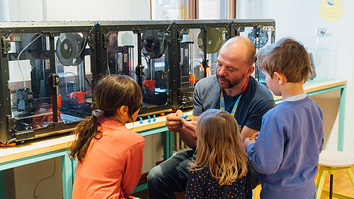 Ein Mann und drei Kinder sitzen vor einer Reihe von 3D-Druckern. Der Mann erklärt etwas, die Kinder haben Spaß und hören zu.