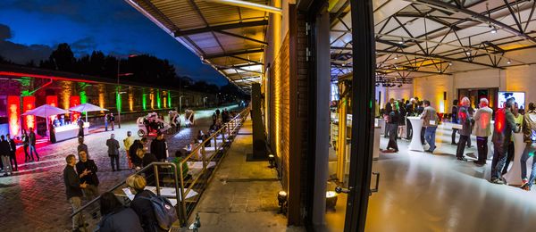In der Ladestraße wird eine Abendveranstaltung mit Straßenfestcharakter gezeigt. Das Bild ist mittig geteilt, zeigt auf der rechten Seite einen Blick in die Halle 5 und auf der linken Seite einen Blick in den farbig beleuchteten Außenbereich.