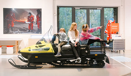 Drei Kinder sitzen in einer Ausstellungshalle auf einem schwarz-gelben Schneemobil.