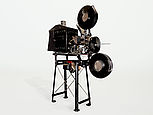 Der Filmprojektor Ernemann I ist auf einem Vorführtisch montiert. Oben über dem Objektiv und schräg nach vorne versetzt, unter dem Objektiv, sind zwei Filmrollen angebracht.
