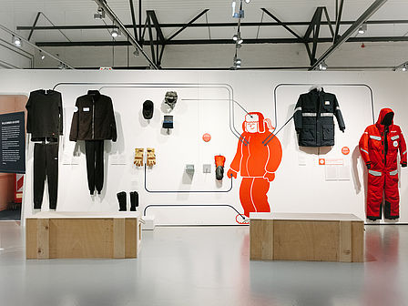 An einer hellen Wand hängen mehrere Kleidungsstücke: dicke Anoraks, ein roter Ganzkörperanzug, Handschuhe und Mützen.