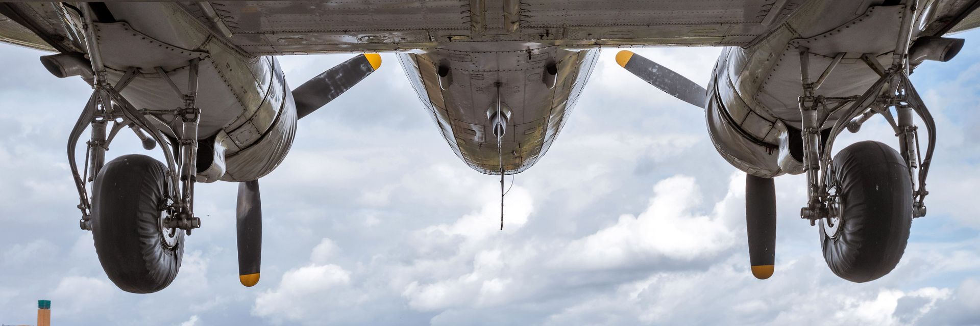 Untersicht auf ein Flugzeug, darunter blickt man auf einen bewölkten Himmel und die Dächer einer Stadt.