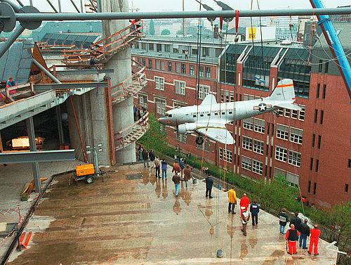 Auf einer Betonterrasse eines Rohbaus stehen viele Menschen, während zugleich ein Flugzeug mittels eines Krans auf die Ebene herabgelassen wird.