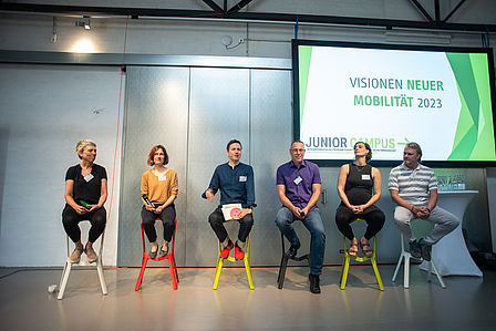 Sechs Personen sitzen nebeneinander auf einer Bühne und blicken in den Raum. Im Hintergrund wird die Präsentation des Junior Campus zu Visionen neuer Mobilität 2023 auf Leinwand gezeigt.