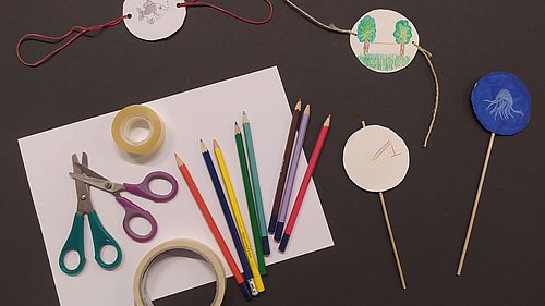Ein weißes Blatt Papier, darauf Scheren, Klebeband und Bunstifte. Daneben liegen die fertig gebastelten Thaumatrope auf dem schwarzen Untergrund.