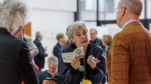 Drei Personen stehen während einer Veranstaltung an einem Stehtisch und disutieren. Eine Dame hält ein Informationsheft zum Thema Zukunft in der Hand.