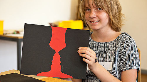 Ein junges Mädchen hält ein Blatt Papier mit schwarzen und roten Formen vor sich. Der rote Bereich in der Mitte sieht aus wie eine Vase. Die schwarzen Bereiche links und rechts wie zwei Gesichter im Profil, die sich anschauen.
