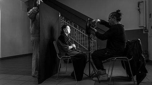 Schwarz-Weiß-Fotografie. Eine Person sitzt auf einem Stuhl. Hinter ihr hält eine weitere Person eine schwarze Leinwand hoch. Vor ihr sitzt eine weitere Person auf einem Stuhl und fotografiert die erste Person.