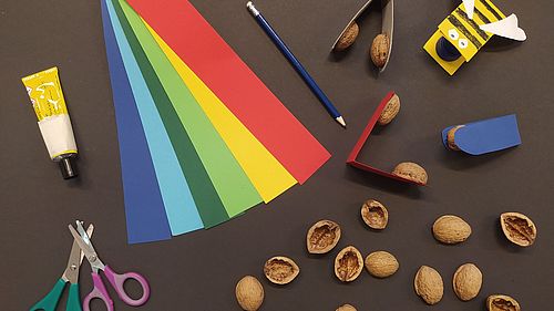 Scheren, eine Tube Klebstoff, bunte Papierstreifen und Walnussschalen liegen auf schwarzem Untergrund. Rechts oben liegen die fertigen klappernden Walnüsse.