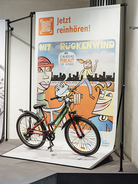 Fahrrad-Ausstellung mit Banner des Podcasts „Mit Rückenwind – Der Fahrrad Podcast für Kinder“. Buntes Fahrrad vor Plakat mit Zeichnung: Figuren, Stadtsilhouette und Slogan „Jetzt reinhören!“ mit QR-Code.