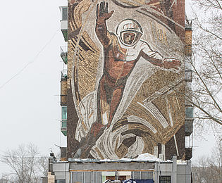 A car and a truck bearing a Coca-Cola advertisement are driving along a snow-covered road. In the background is a high-rise building with a mosaic mural depicting a larger-than-life cosmonaut floating up to the sky.