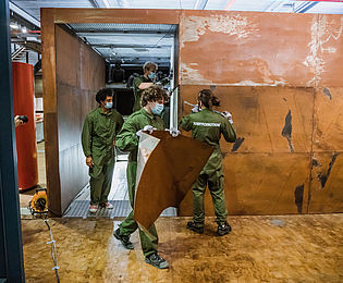 Mehrere Menschen in grünen Arbeitsanzügen transportieren Materialien aus einem braunen Metallkubus.