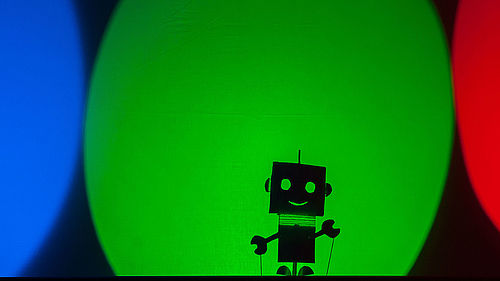 Auf einen ebenen Hintergrund scheint kreisförmig grünes Licht. Der obere und untere Teil des Kreises sind abgeschnitten. Unten, etwa mittig, befindet sich der Schatten einer kleinen Roboter-Figur. Links neben dem grünen befindet sich ein blauer und rechts ein roter Lichtfleck. 