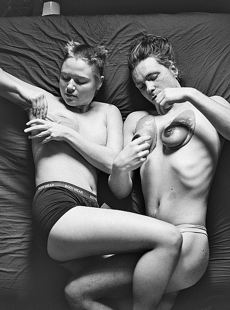 Das schwarz-weiße Foto zeigt zwei Personen in Unterwäsche, die eng nebeneinander auf einem Bett liegen. Die linke Person, offenbar eine Frau mit kurzen Haaren, trägt Boxershorts und verdeckt mit dem Arm ihre Brust. Die rechte Person, wohl ein Mann mit halblangem Haar, trägt einen Tanga. Auf dem Brustkorb sind zwei Plastikobjekte positioniert, die wie weibliche Brüste aussehen.