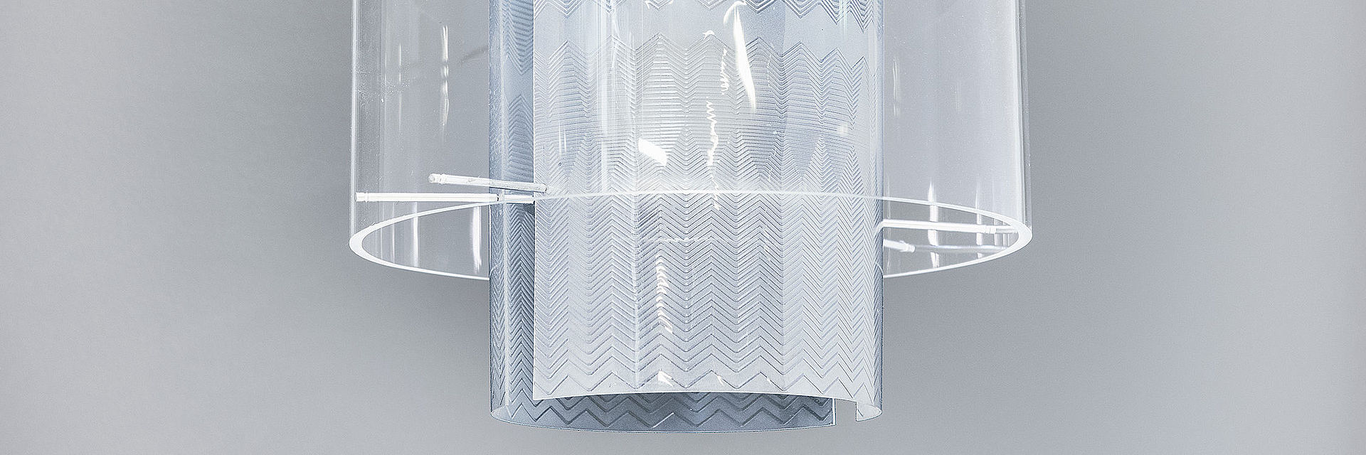 Die Lampe hängt an einem weißen Kabel. Die Glühbirne ist von durchsichtigen, rund gebogenen Plexiglasscheiben umschlossen, die mit eingravierten Zickzacklinien verziert sind.