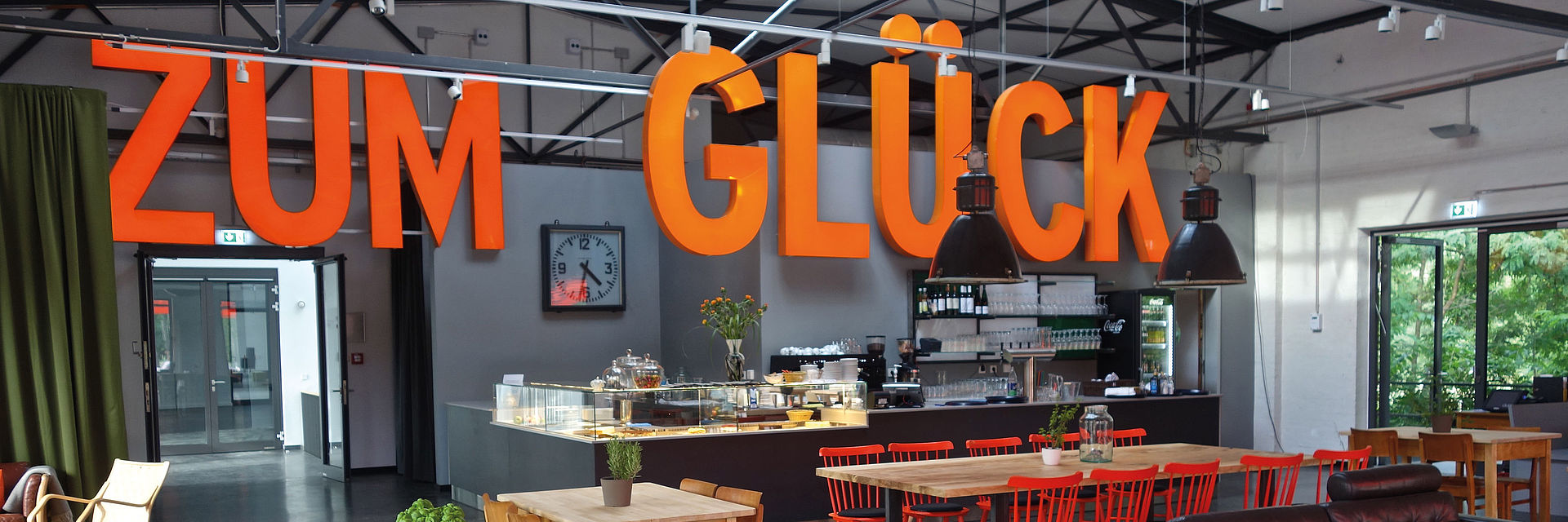 Blick in den Gastraum eines Cafés mit Tischen und Stühlen, von der Decke hängt ein orangener Schriftzug „Zum Glück“.