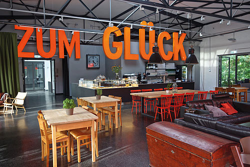 Blick in den Gastraum eines Cafés mit Tischen und Stühlen, von der Decke hängt ein orangener Schriftzug „Zum Glück“.