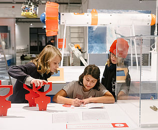 Drei Kinder messen an einem Ausstellungstisch die Dicke einer "Eisscholle" und tragen die Ergebnisse in ein Forschungsheft ein.
