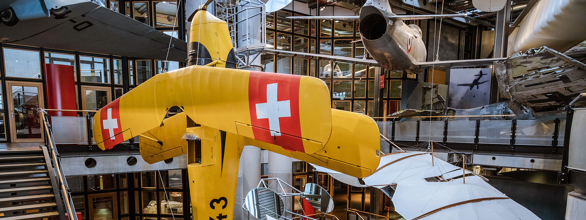 Ein großes gelbes Flugzeug hängt senkrecht im offenen Treppenhaus des Neubaus. Im Hintergrund hängen weitere Flugzeuge und Flugapparate. 