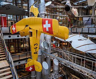 Ein großes gelbes Flugzeug hängt senkrecht im offenen Treppenhaus des Neubaus. Im Hintergrund hängen weitere Flugzeuge und Flugapparate. 