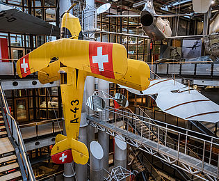 Ein großes gelbes Flugzeug hängt senkrecht im offenen Treppenhaus des Neubaus. Im Hintergrund hängen weitere Flugzeuge und Flugapparate. 