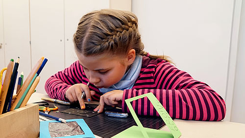 Ein Mädchen sitzt an einem Tisch und bastelt mit dunklem Papier auf einer dunklen Unterlage. Auf dem Tisch steht eine Box mit Buntstiften. Daneben liegen zwei Karten aus Buntpapier. Bei einer davon ist aus der Vorderseite ein Viereck wie ein Sichtfenster ausgeschnitten.