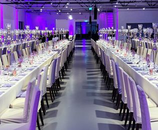 Die Hallen 5 und 6 der Ladestraße sind für ein Dinner an langen Tafeln hergerichtet. Tischwäsche und Hussen sind in Weiß gehalten. Die Tische sind eingedeckt und mit Kerzen und Blüten dekoriert.