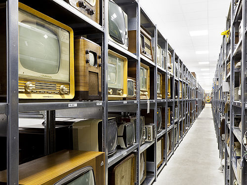 Ein schmaler Gang zwischen langen Metallregalen. In den Regalen stehen viele historische Fernsehgeräte mit Holzgehäuse.