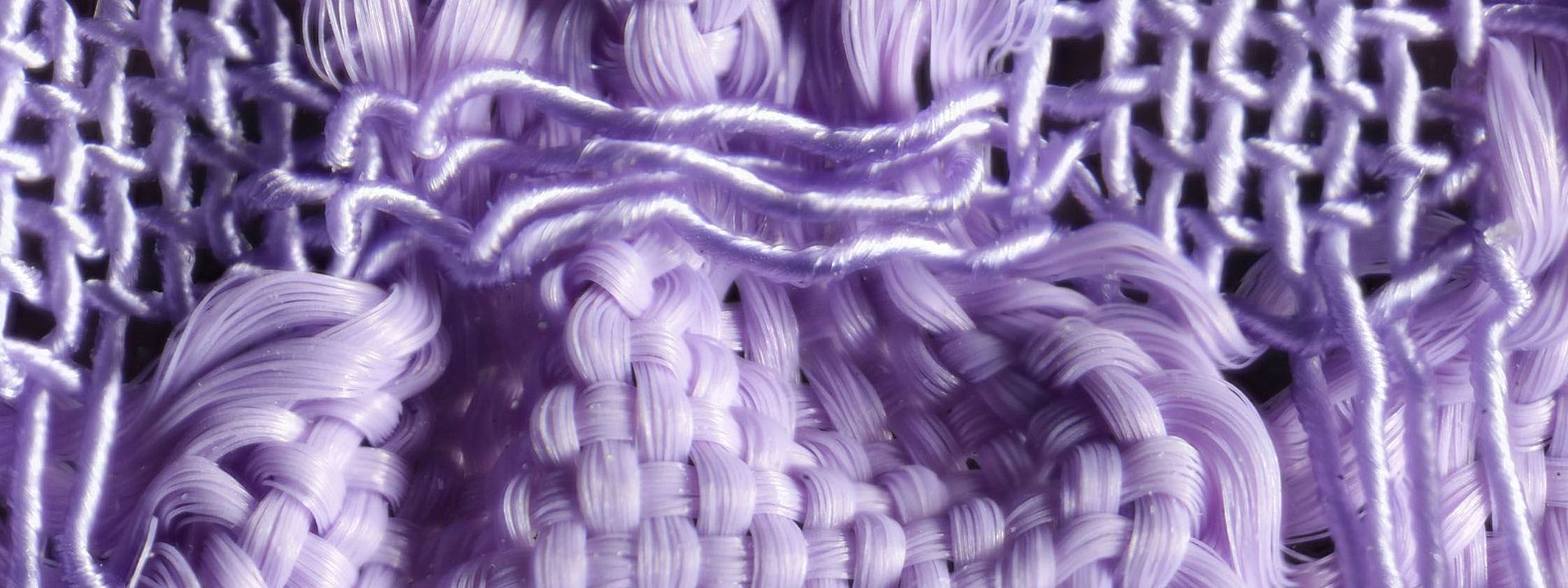 Detailaufnahme eines lilafarbenen Gewebes. Durch zwei unterschiedlich elastische Garne, Gummilitze und Seide, tritt das Blasenmuster des Stoffes besonders hervor. 