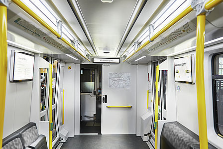Blick in den Innenraum eines U-Bahnwagens. Der Eindruck ist hell und groß. Die Sitze sind blau-schwarz gemustert, die Haltestangen sind gelb.