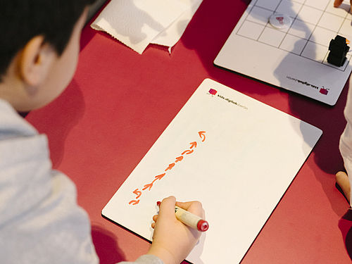 Ein KInd zeichnet mit einem orangfarbenen Stift Pfeilbehle auf ein Blatt Papier.