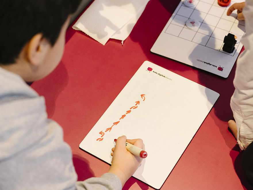 Ein KInd zeichnet mit einem orangfarbenen Stift Pfeilbehle auf ein Blatt Papier.