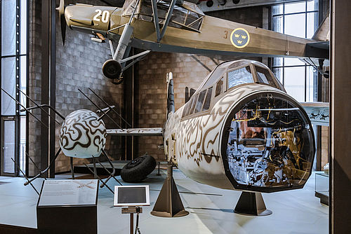 Frontaler Blick auf ein Kampfflugzeug. Die Nase des Flugzeugs ist abmontiert, so dass man in das beleuchtete Innere blicken kann. Im Hintergrund hängt ein weiteres Flugzeug.