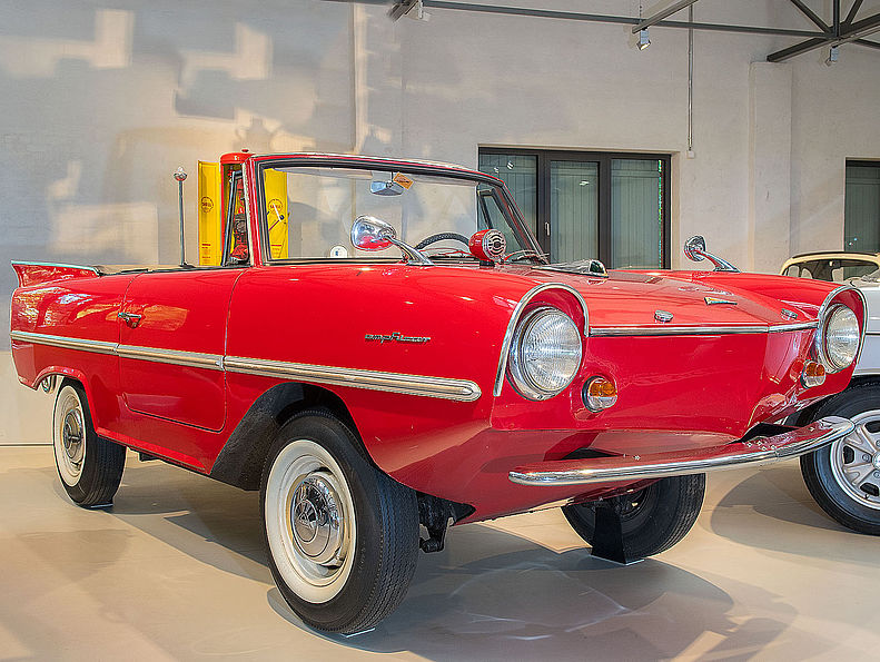 Das feuerrote Amphicar hat deutlich mehr Bodenfreiheit als ein normales Auto. Es sieht aus wie ein Cabrio und ähnelt im Design anderen Wagen der frühen 1960er Jahre: runde Glubschaugen-Scheinwerfer, angedeutete Flossen am Heck.