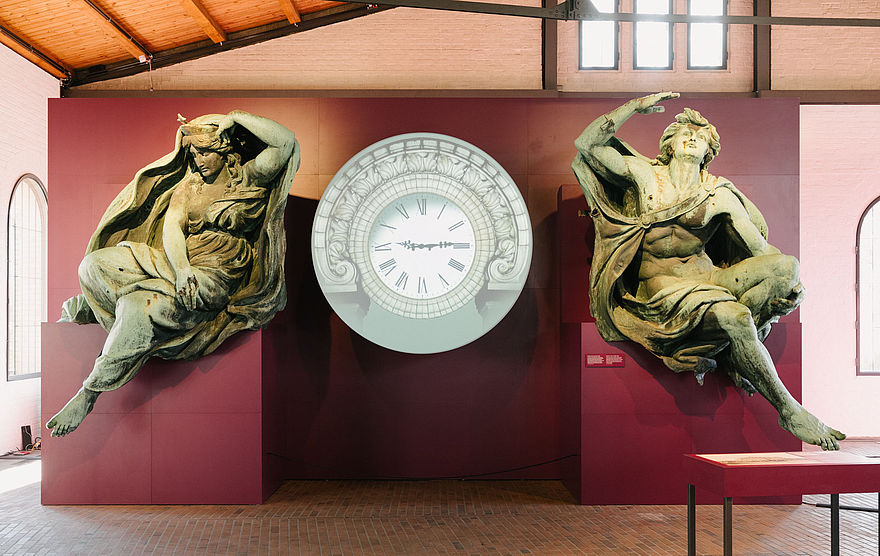 Zwei große, grün angelaufene Bronze Figuren stehen links und rechts. Zwischen Ihnen steht eine große Uhr.