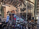 Eine Fahrradwerkstatt mit zahlreichen aufgereihten Fahrrädern und Ersatzteilen. Fahrräder und Felgen hängen an Gestellen und von der Decke, umgeben von Werkzeugen, Regalen und einer Leiter.