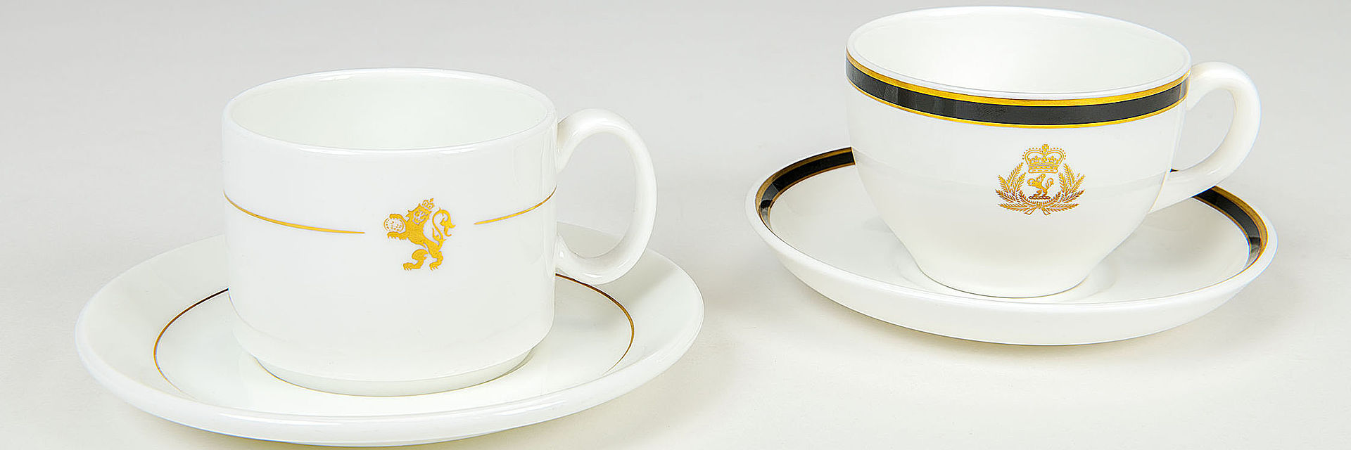 Auf dem Foto sind zwei weiße Porzellantassen abgebildet. Die linke Tasse ist zylinderförmig, mit goldenem Streifen und einem goldenen Löwen als Symbol der Reederei Cunard. Die rechte Tasse ist bauchiger geformt, hat einen gold-schwarzen Rand und trägt eine prunkvollere Version des Reederei-Logos; sie wirkt insgesamt edler.
