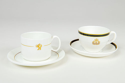 Auf dem Foto sind zwei weiße Porzellantassen abgebildet. Die linke Tasse ist zylinderförmig, mit goldenem Streifen und einem goldenen Löwen als Symbol der Reederei Cunard. Die rechte Tasse ist bauchiger geformt, hat einen gold-schwarzen Rand und trägt eine prunkvollere Version des Reederei-Logos; sie wirkt insgesamt edler.