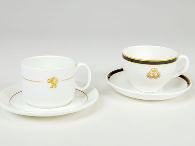 Auf dem Foto sind zwei weiße Porzellantassen abgebildet. Die linke Tasse ist zylinderförmig, mit goldenem Streifen und einem goldenen Löwen als Symbol der Reederei Cunard. Die rechte Tasse ist bauchiger geformt, hat einen gold-schwarzen Rand und trägt eine prunkvollere Version des Reederei-Logos; sie wirkt insgesamt edler.