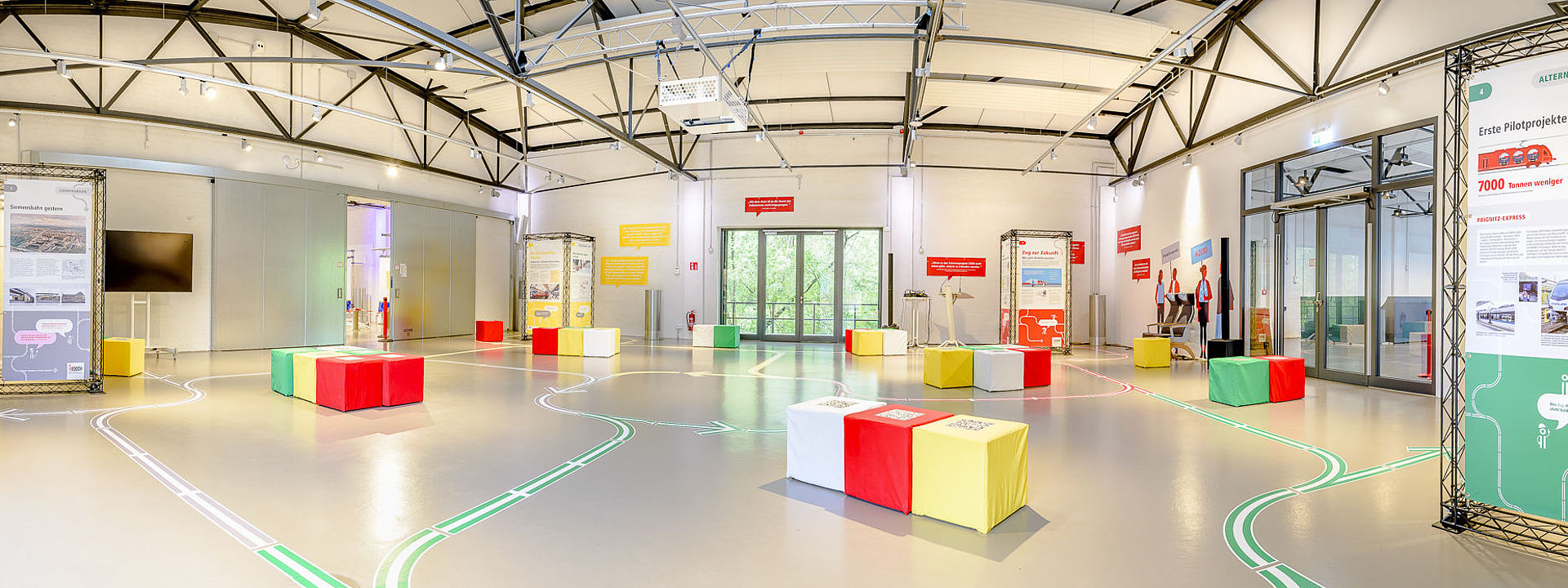 Blick in die Ausstellung, rechts Stellwände mit Text, bunte Hocker, Bodenmarkierung mit geschwungenen Pfeilen.