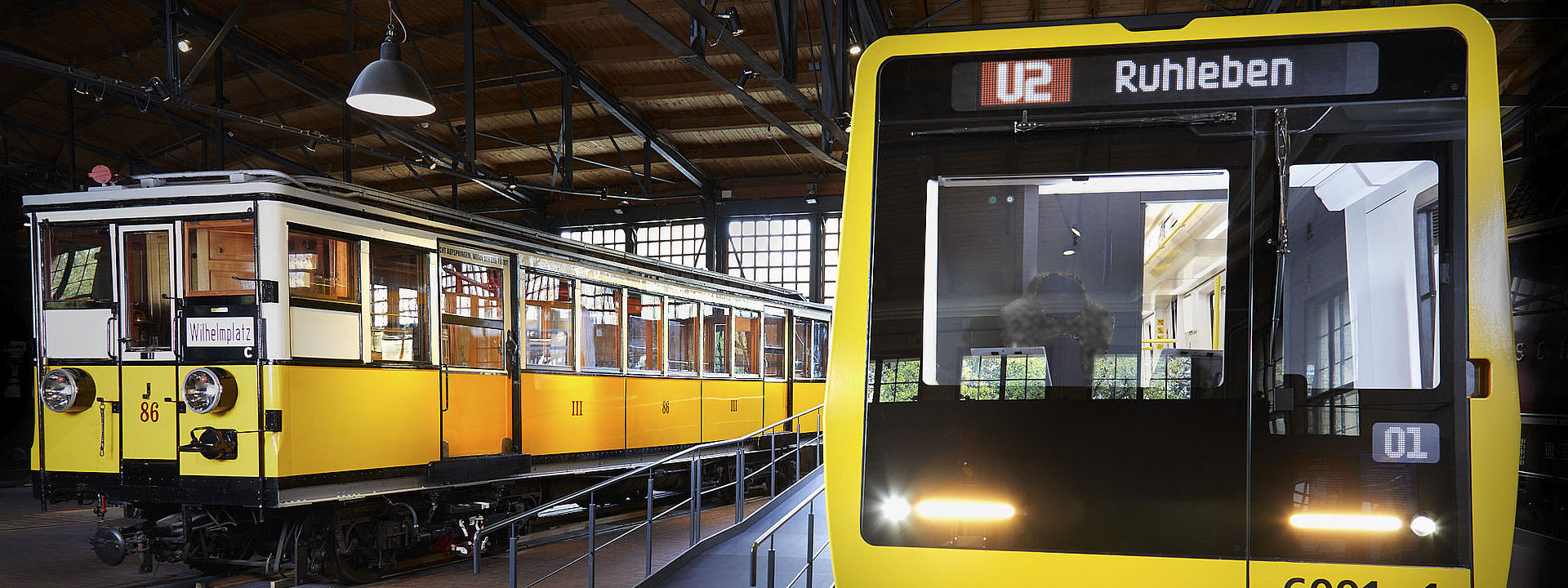 Blick auf zwei nebeneinander stehende U-Bahnwagen. Beide sind gelb. Der Wagen rechts sieht sehr modern aus, der Wagen links ist ein historisches Fahrzeug.